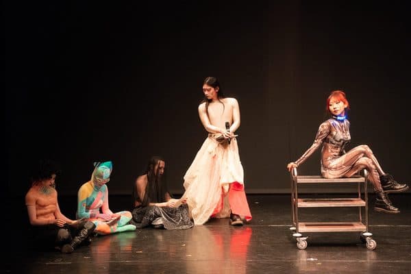 Performers in vivid body paint and costume on a black-box stage.