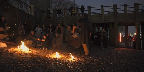 A crowd gathered around small fires on a Thames foreshore at dusk.